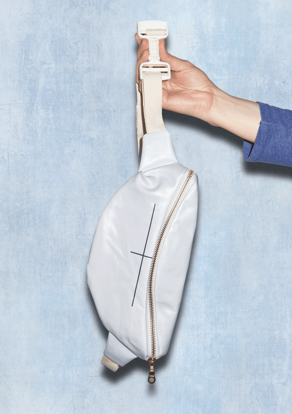 White waist bag with a gold zipper being held against a light blue background