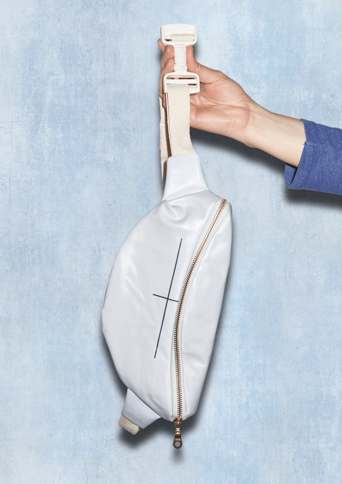 White waist bag with a gold zipper being held against a light blue background