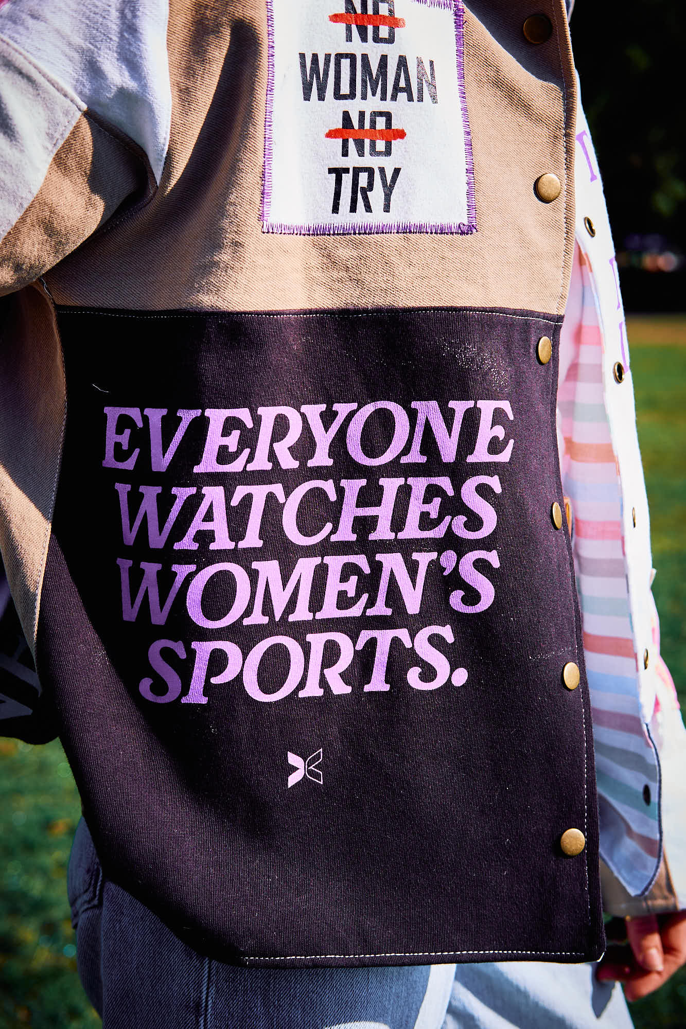 Person wearing a jacket with text 'Everyone watches women's sports' and a patch above it with text 'No Woman No Try'.