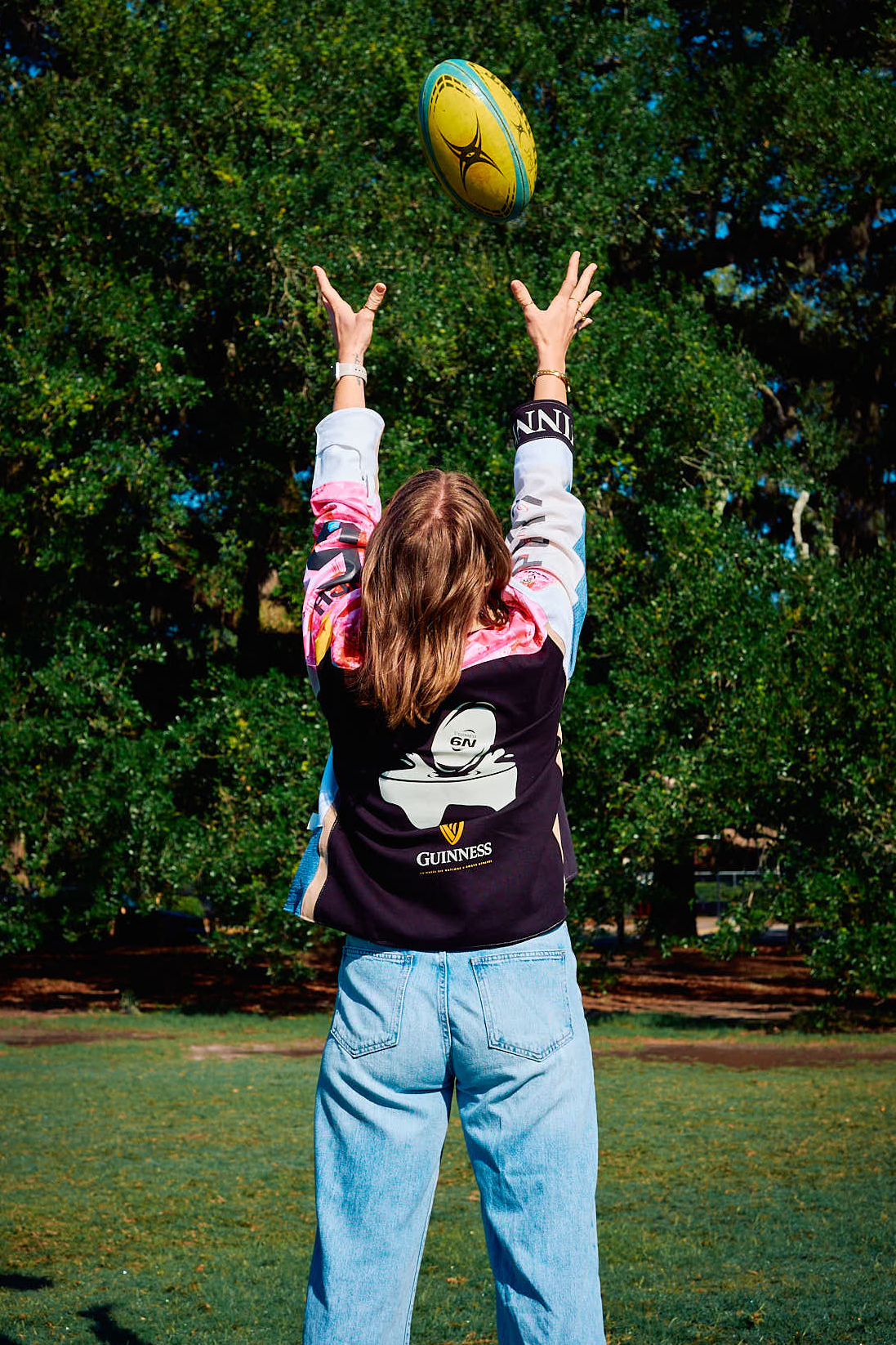 Person in a black jacket with a Guinness Six Nation Rugby design reaching up to catch a Rugby ball in an outdoor setting with trees in the background