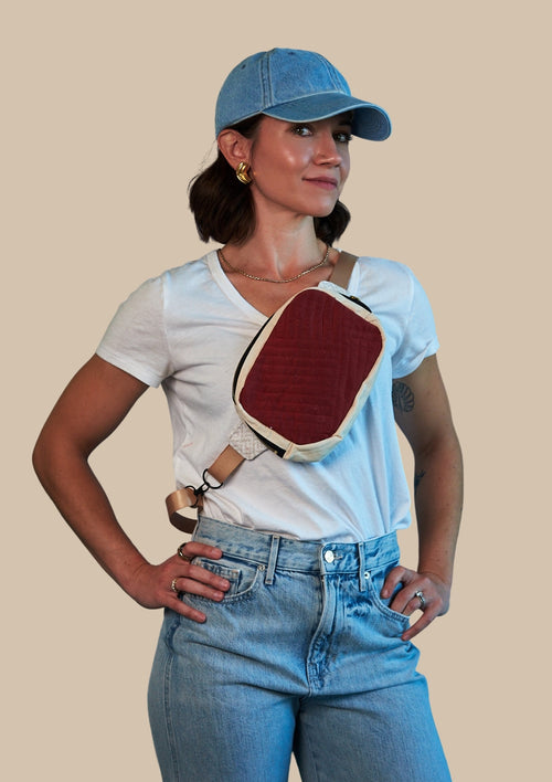 Person wearing a white t-shirt, blue jeans, and a blue cap with a red and beige bag over one shoulder upcycled from comforter on a beige background.