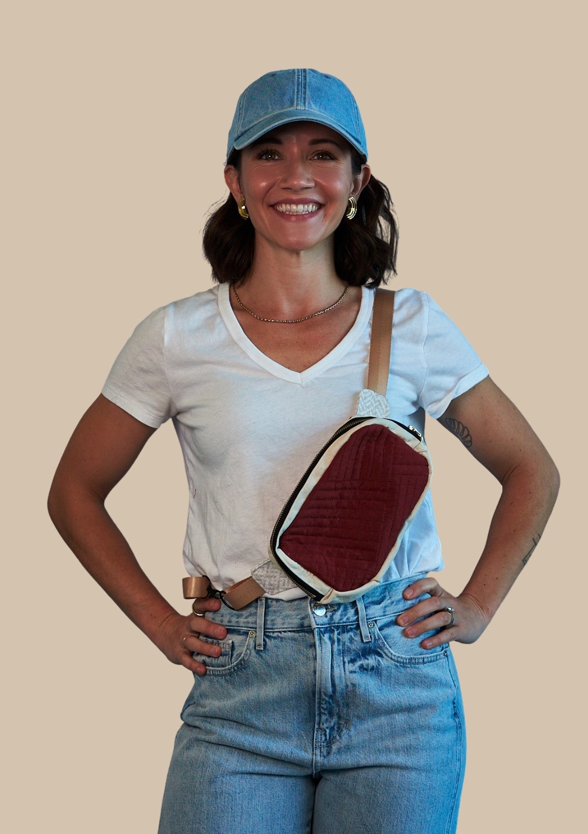 Person wearing a white t-shirt, blue jeans, and a blue cap with a red and beige crossbody bag over one shoulder upcycled from comforter on a beige background.