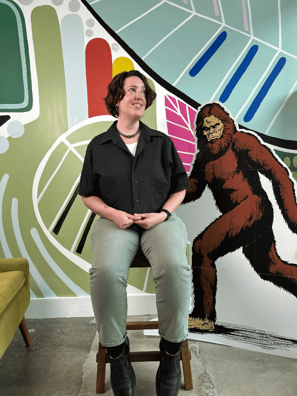 Person wearing a black cropped shirt upcycled from a dress shirt, sitting on a chair in front of a colorful wall with a cartoon-like depiction of a bigfoot.