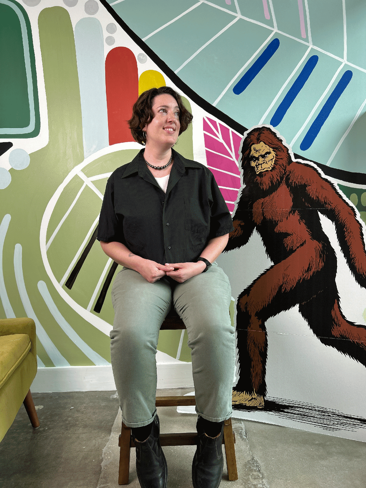 Person wearing a black cropped shirt upcycled from a dress shirt, sitting on a chair in front of a colorful wall with a cartoon-like depiction of a bigfoot.