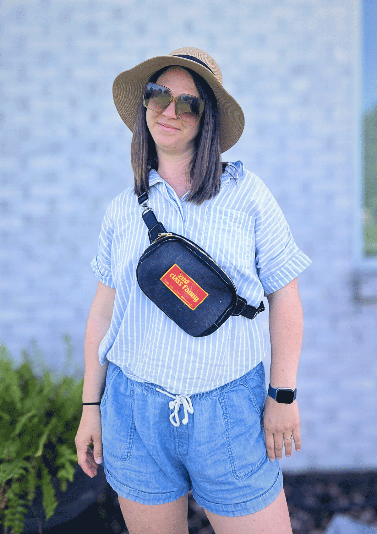 Person wearing a striped shirt, shorts, and a hat with a crossbody bag with a pink and yellow label reads 2nd Class Fanny against a brick wall.
