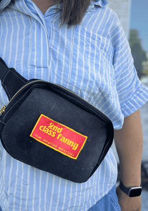 Person wearing a black fanny pack  upcycled from black denim jeans with a pink and yellow label, reads 2nd Class Fanny, standing outdoors.