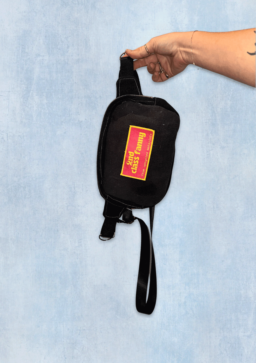 Black bag upcycled from jeans with a pink and yellow label reads 2nd Class Fanny held by a hand on a light blue background