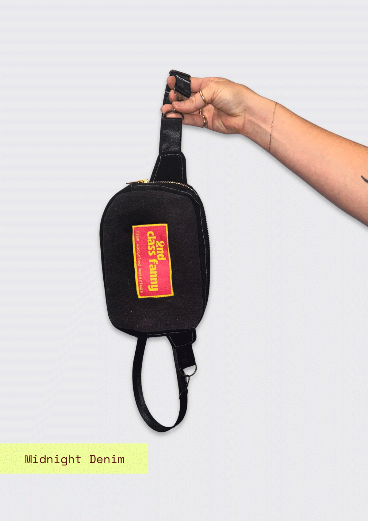 Black waist bag with a colorful label reads 2nd Class Fanny held by a hand on a light gray background