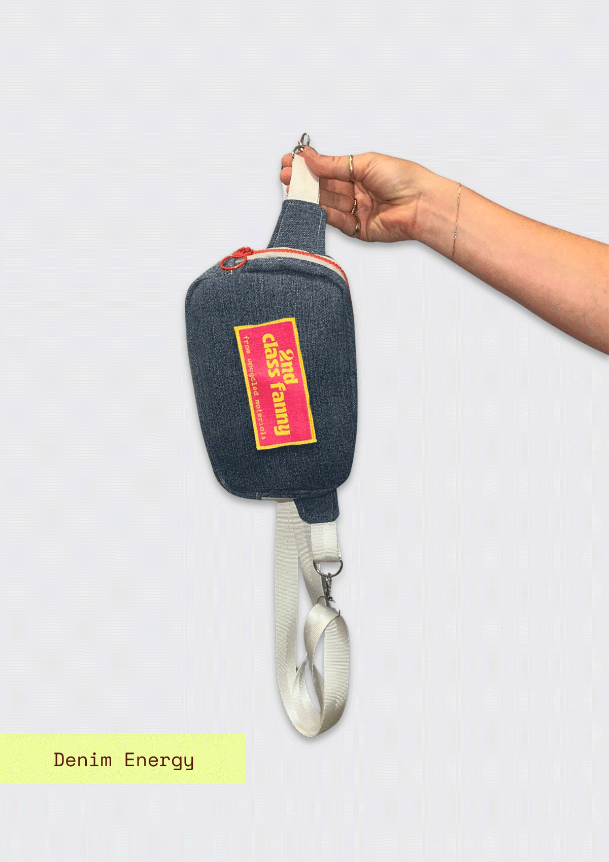 Hand holding Denim fanny pack / cross body bag with label reads 2nd Class Fanny on a gray background