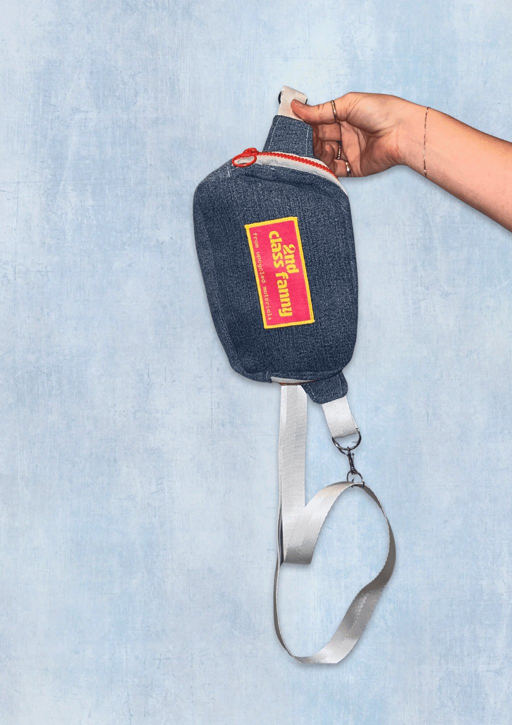 Denim fanny pack or crossbody bag with a brand label held by a hand on a light blue background