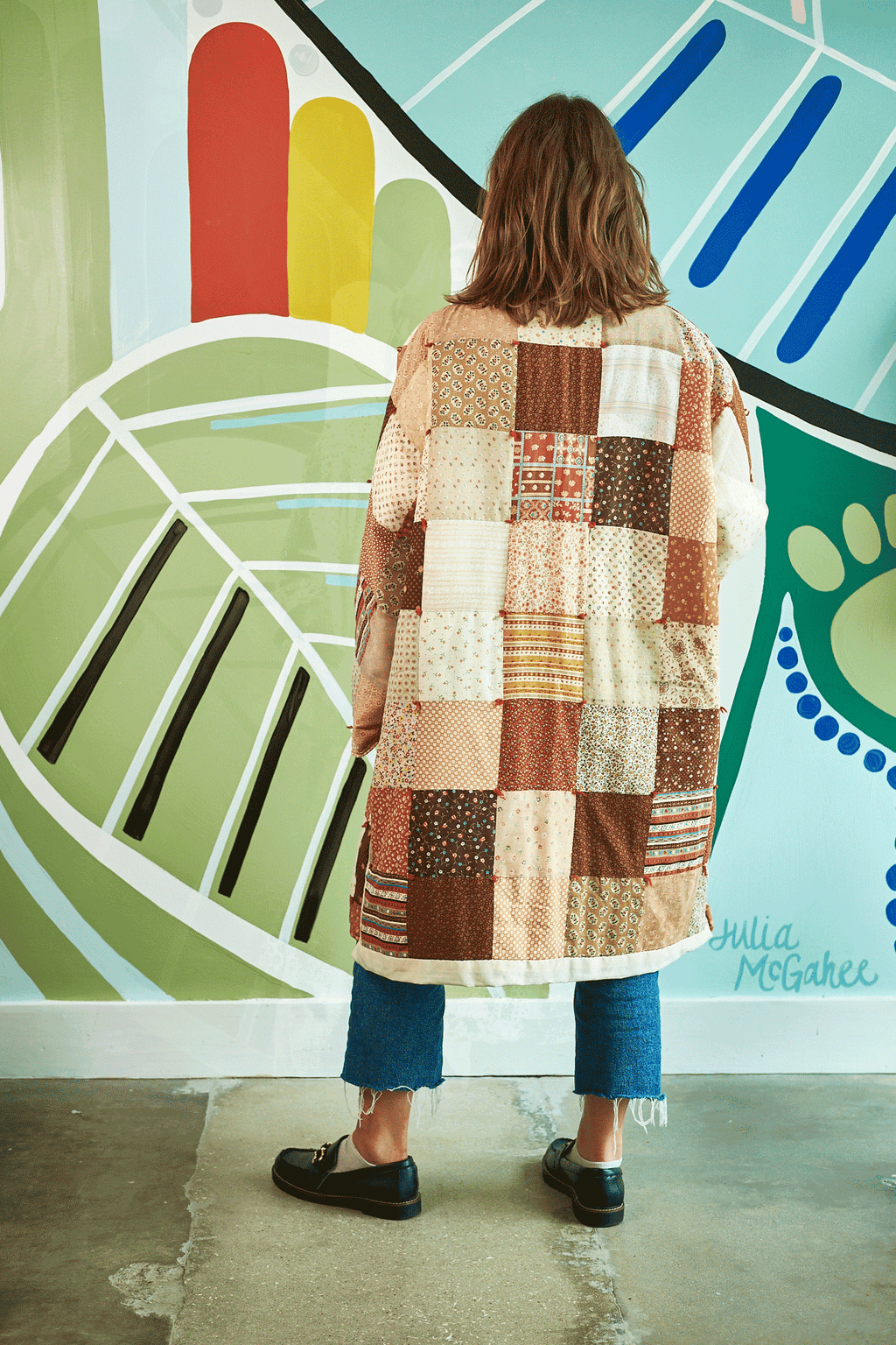 Person wearing a patchwork coat standing in front of a colorful mural.