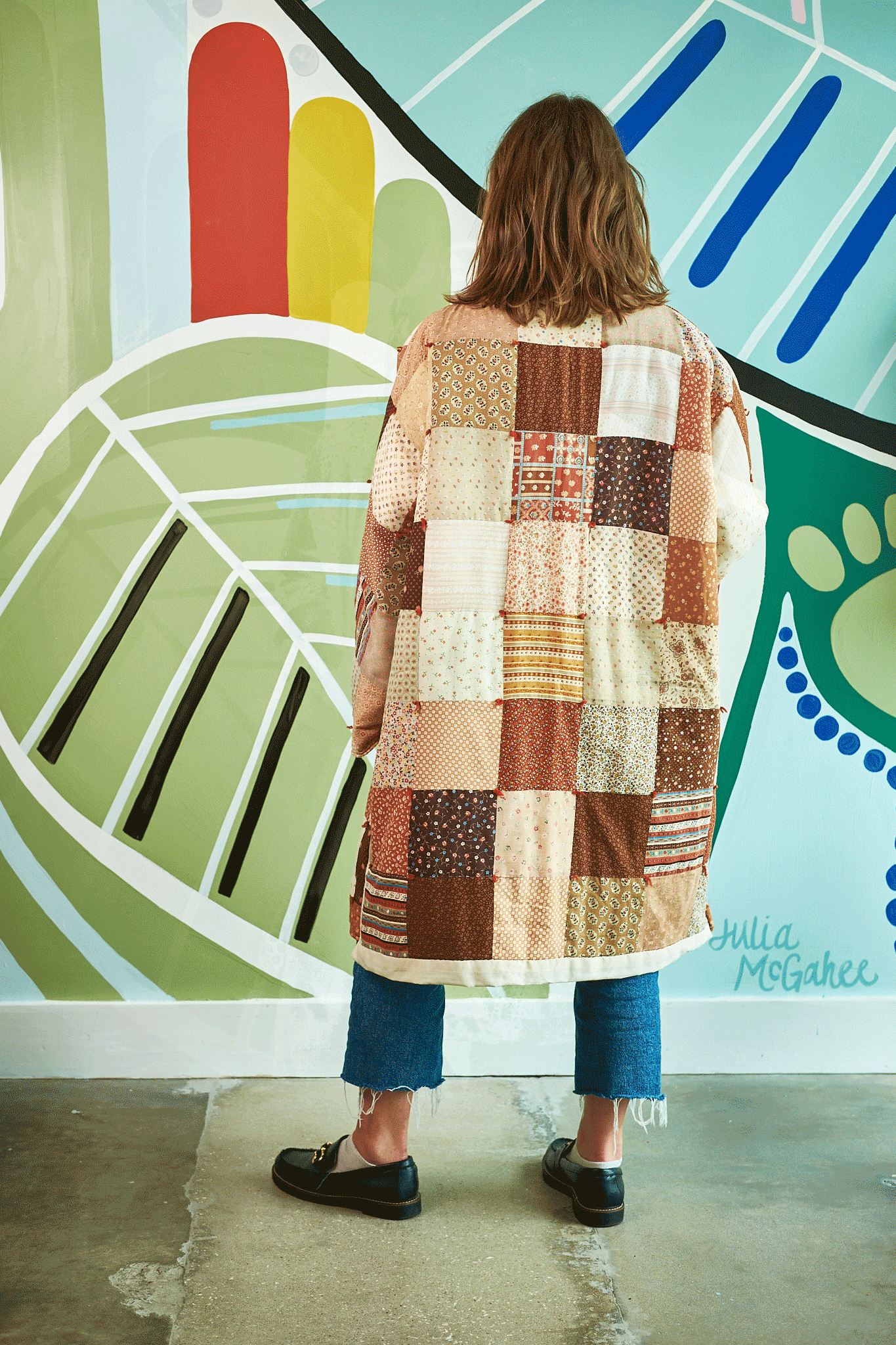Person wearing a patchwork coat standing in front of a colorful mural.