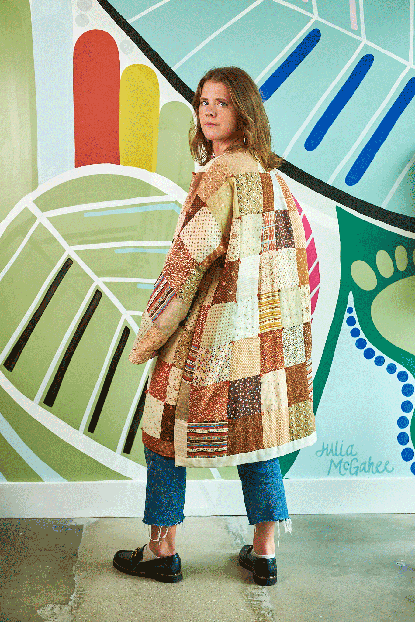 Person wearing a patchwork coat in front of a colorful abstract mural.
