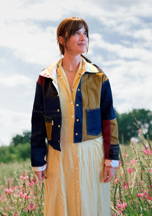 Woman wearing a colorful jacket upcycled from pants over a long dress in a field with pink flowers and a blue sky.