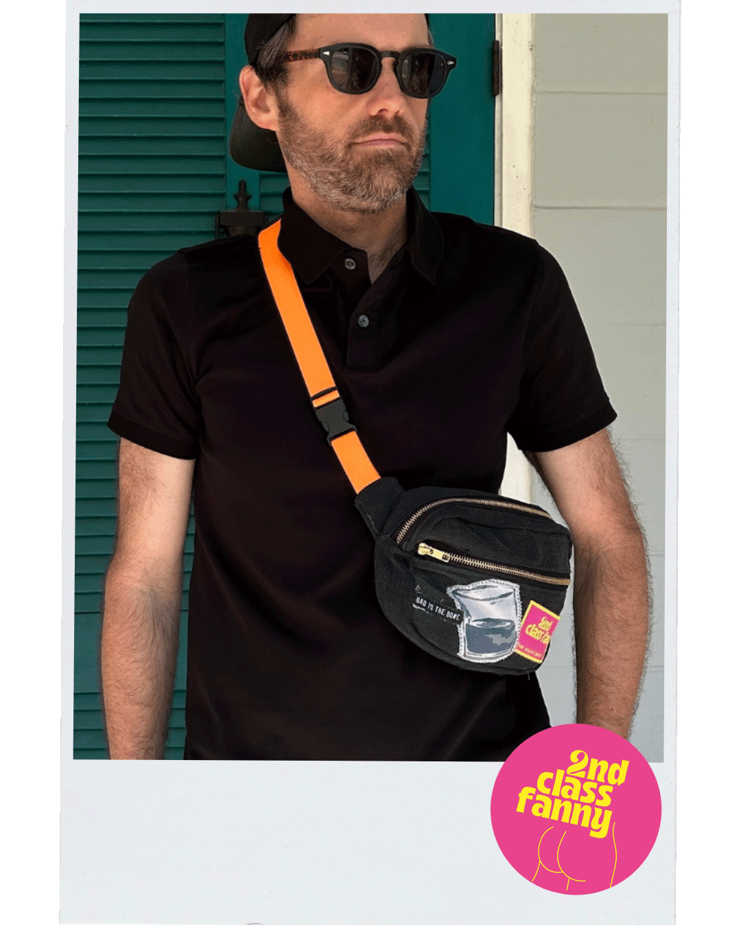 Man wearing a black polo shirt with an orange strap, holding a black fanny pack with a pink label, reads 2nd Class Fanny against a teal door.
