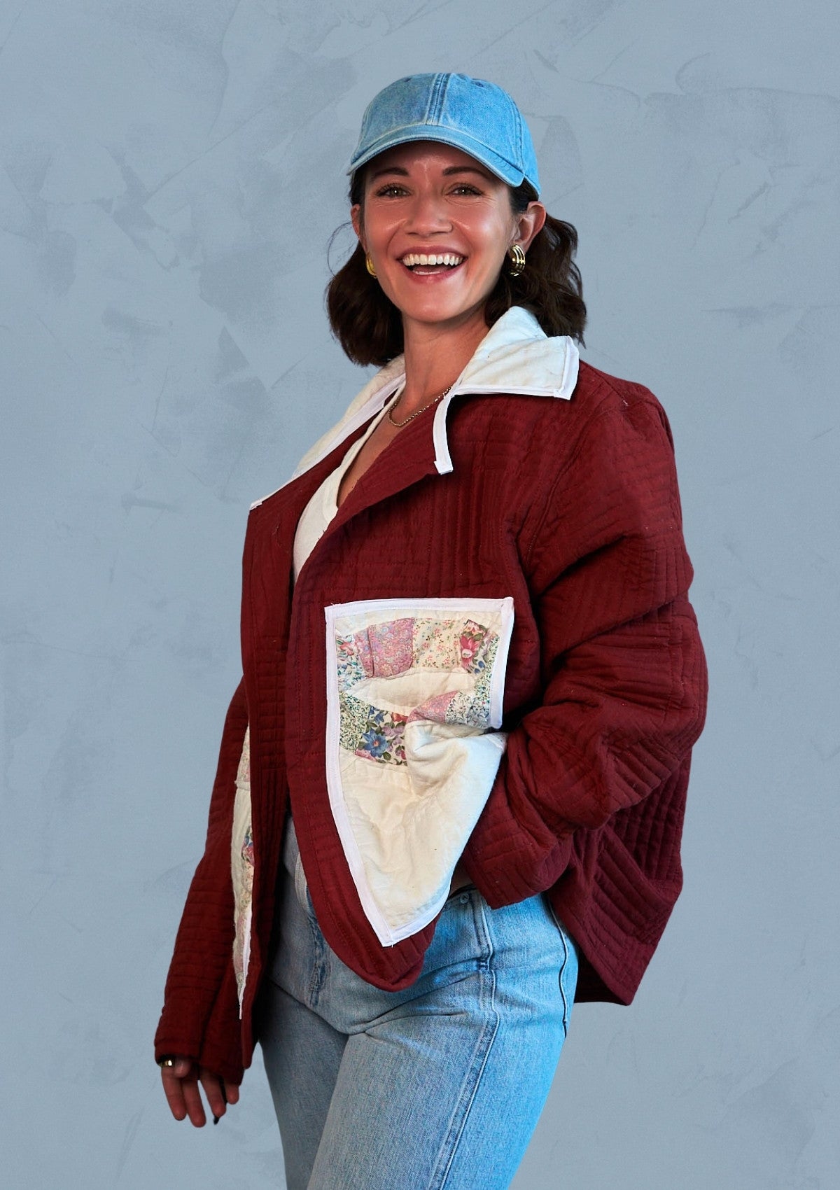 Person wearing a maroon jacket with a patch pockets, upcycled from a quilt blue jeans, and a blue cap against a gray background
