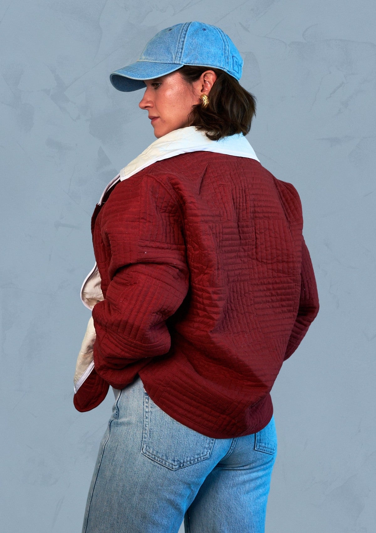 Person wearing a red jacket, upcycled from a quilt, blue jeans, and a blue cap against a gray background