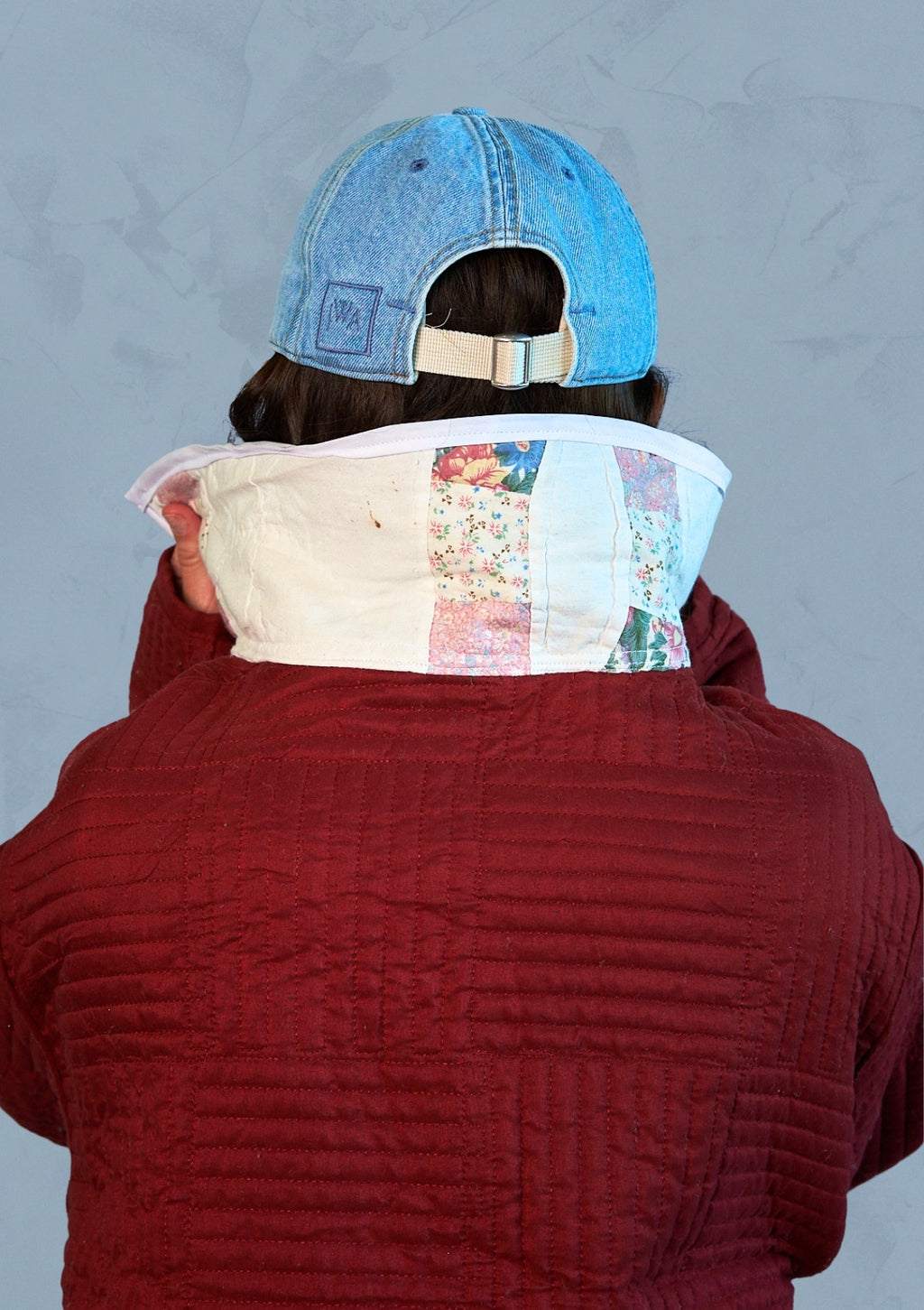 Person wearing a red jacket with a floral patterned collar, upcycled from a quilt  and a blue cap on a gray background