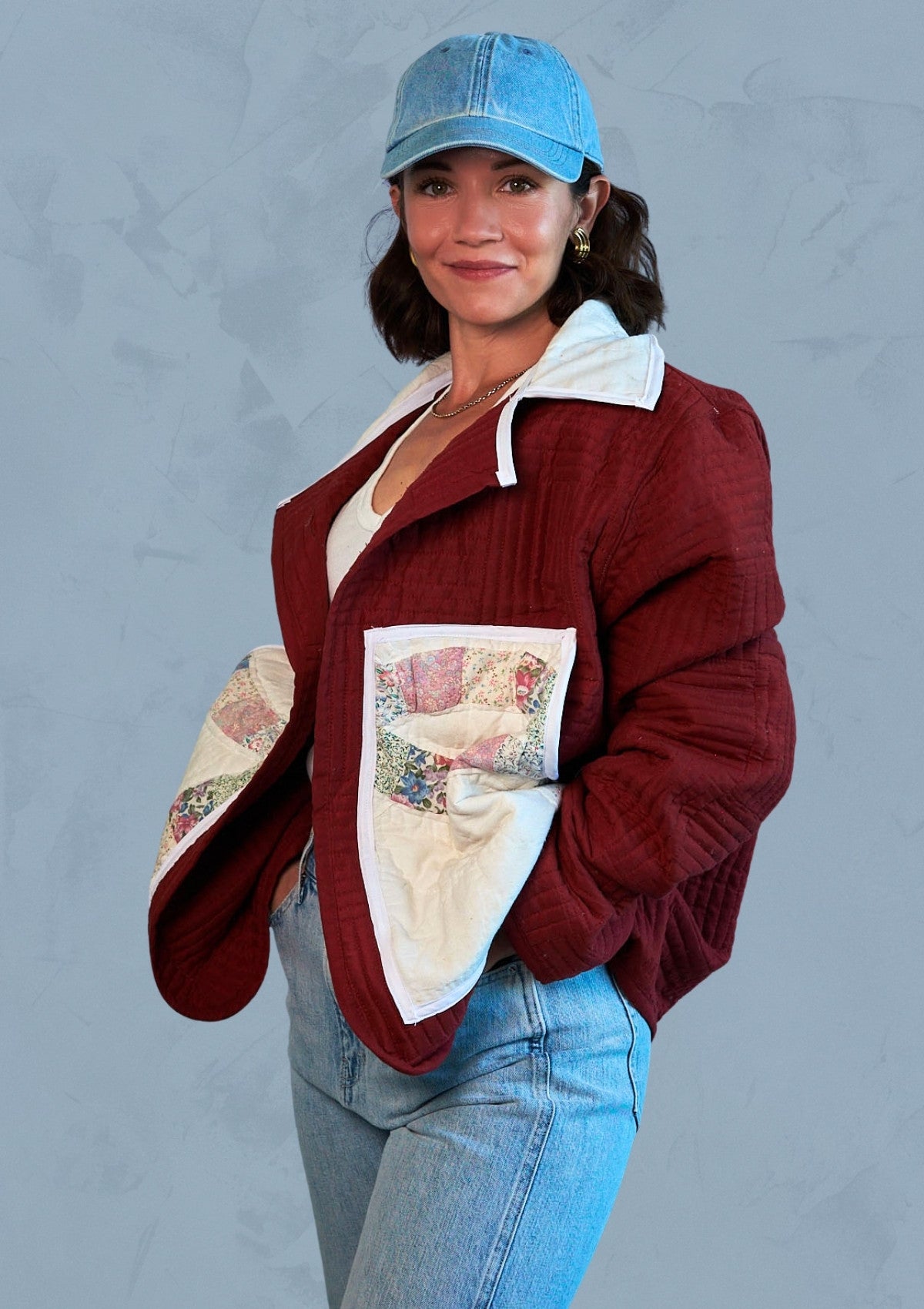 Person wearing a burgundy jacket with patchwork design upcycled from a quilt and blue cap against a gray background