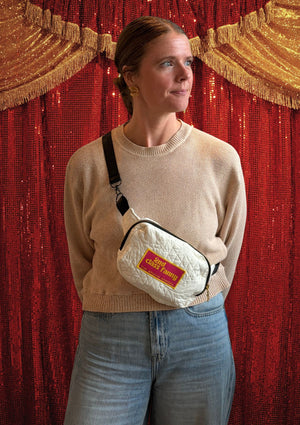 Person wearing a beige sweater and jeans with a white crossbody bag featuring a pink label reads 2nd Class Fanny against a red curtain background
