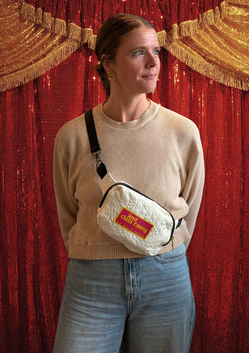 Person wearing a beige sweater and jeans with a white crossbody bag featuring a pink label reads 2nd Class Fanny against a red curtain background