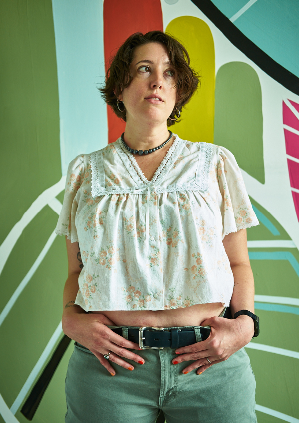 Woman wearing a light floral blouse and green pants against a colorful abstract background