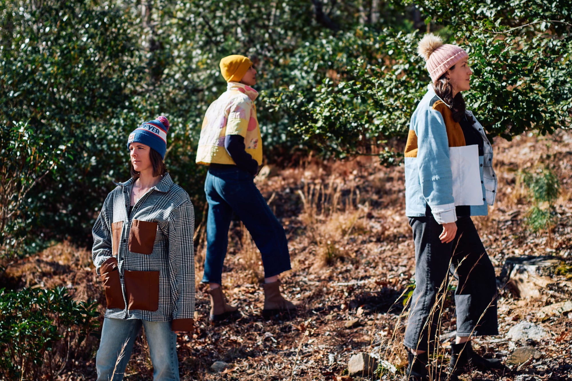 Three people standing outdoors in a natural setting, wearing colorful jackets made from upcycled materials and hats.
