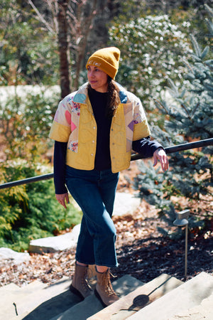 Person wearing a colorful jacket upcycled from quilt and yellow beanie outdoors with trees in the background