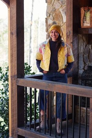 Person wearing a yellow jacket upcycled from quilt and hat on a wooden deck with stone wall background