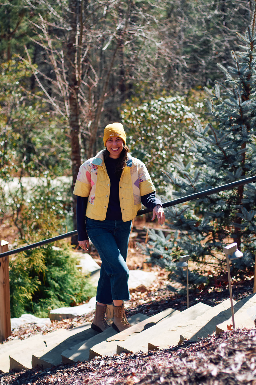 Person in a yellow jacket upcycled from quilt and hat walking on a wooden staircase with trees in the background