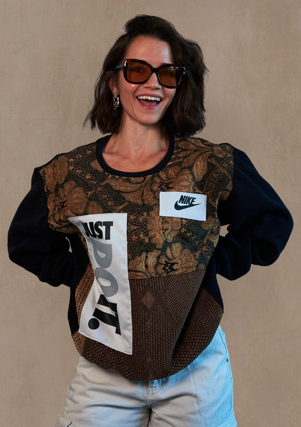 Person in sunglasses wearing a reworked pull over made from upycycled curtains and a Nike just do it tank against a soft muted beige textured background