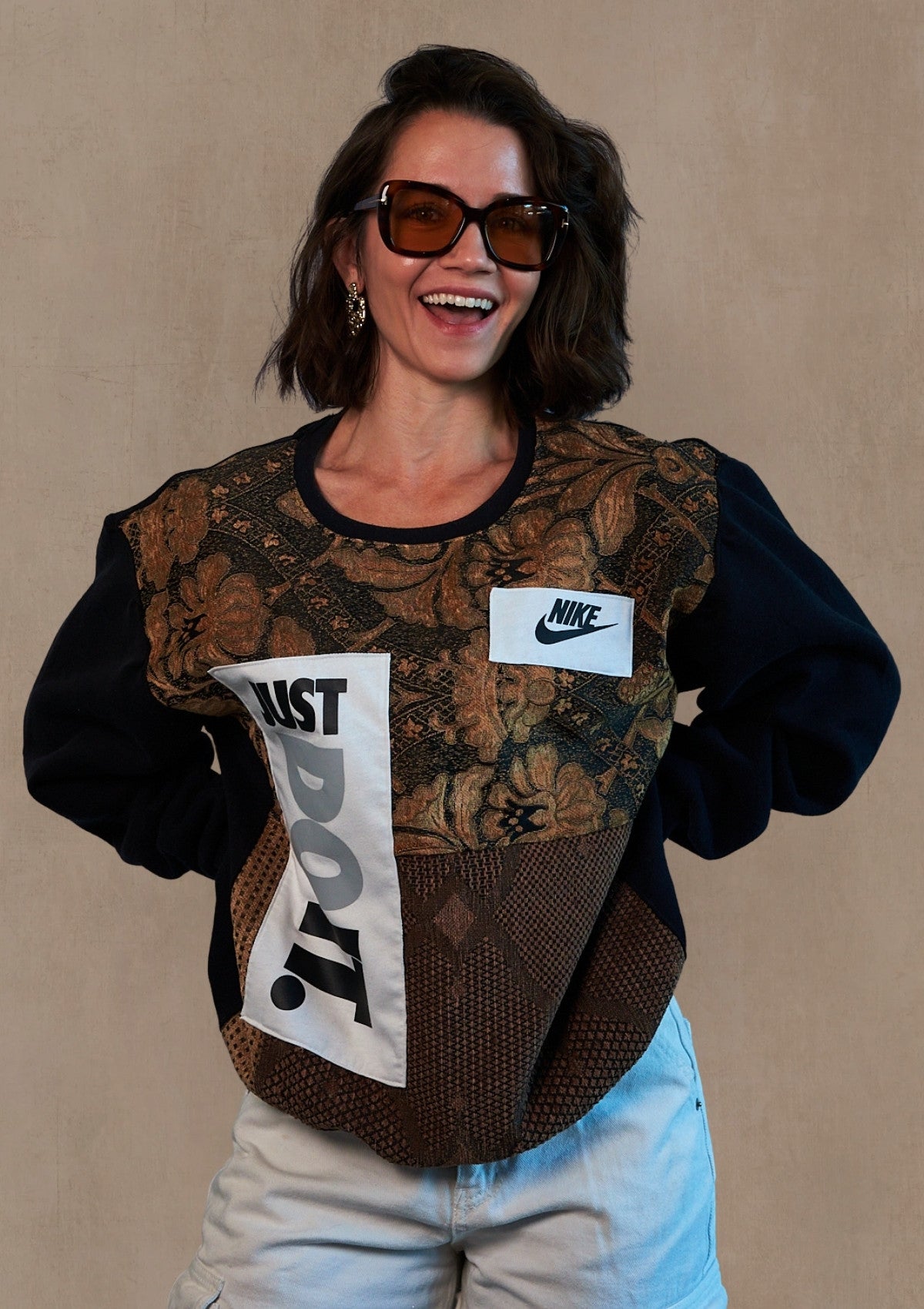 Person in sunglasses wearing a reworked pull over made from upycycled curtains and a Nike just do it tank against a soft muted beige textured background