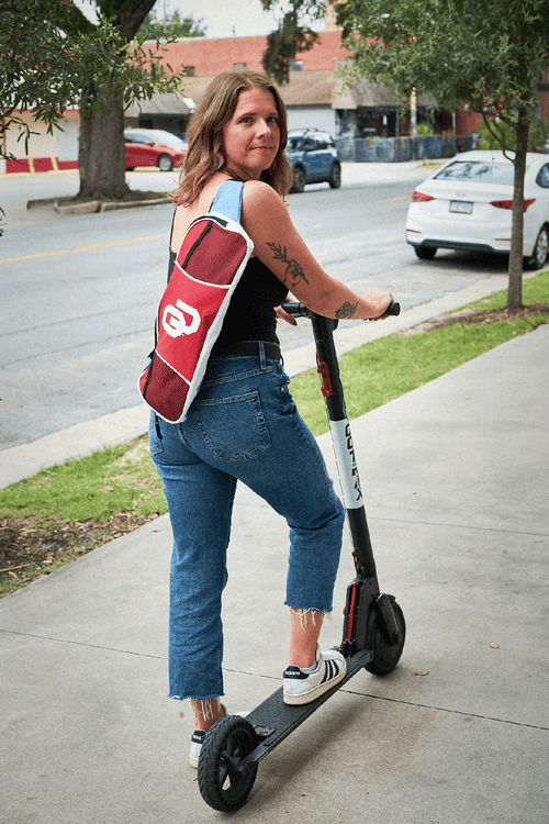 Woman riding a scooter on a sidewalk with a red OU bag over her shoulder.