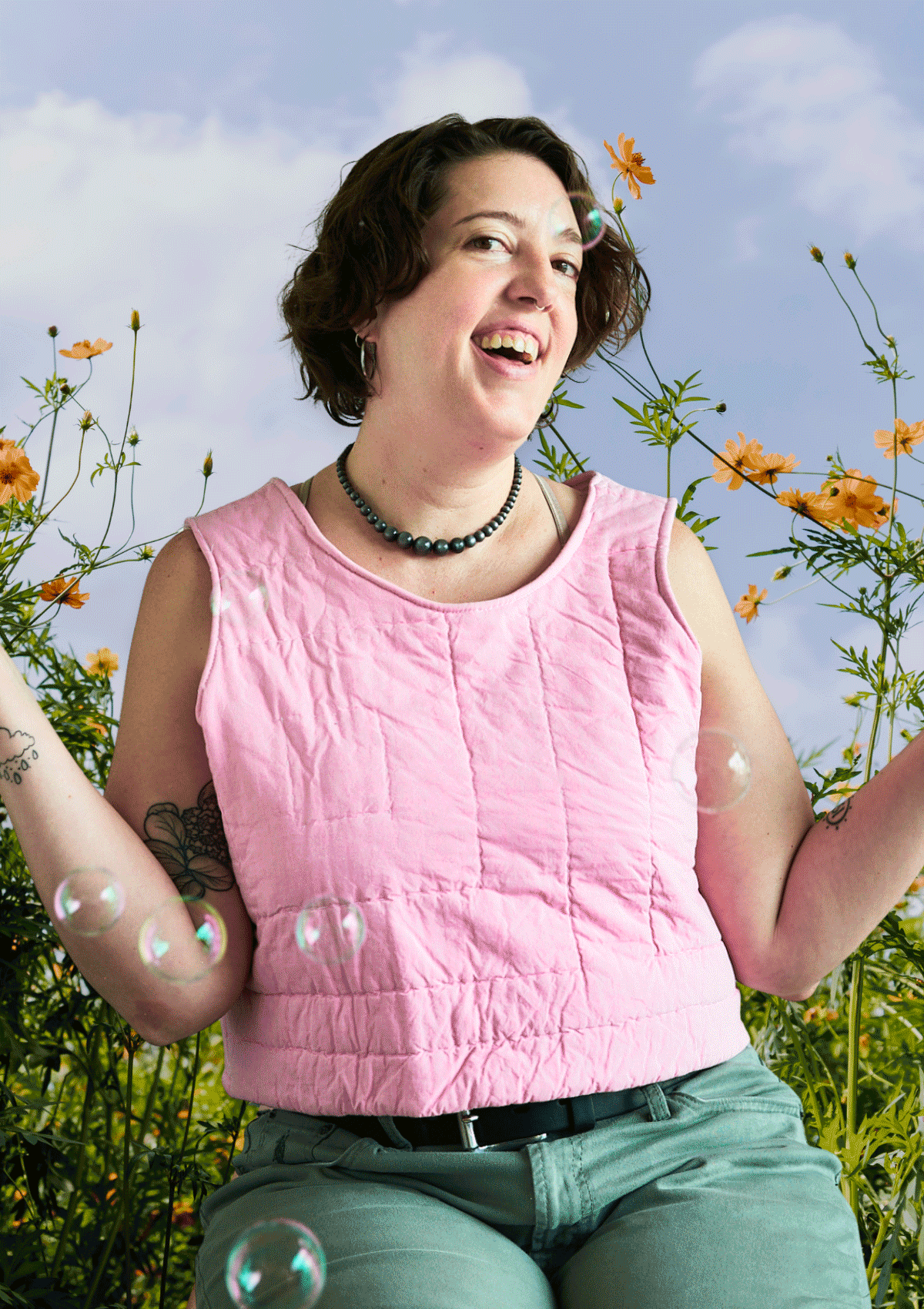 Person wearing a pink sleeveless top upcycled from a quilt and green pants standing among flowers with a blue sky background