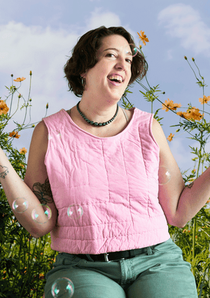 Person wearing a pink sleeveless top upcycled from a quilt and green pants standing among flowers with a blue sky background