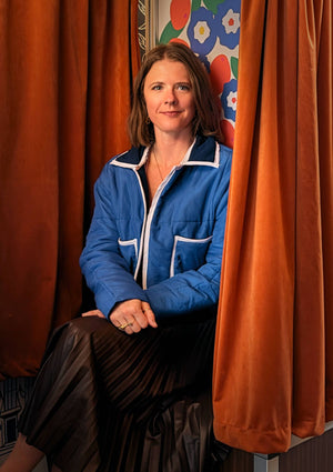 Woman in a blue puffer jacket upcycled from upcycled comforter sitting between orange curtains with a colorful abstract painting in the background.