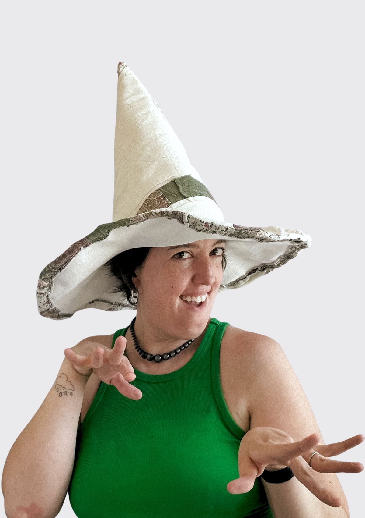 Person wearing a large white witches hat with green trim upcycled from a quilt on a plain background