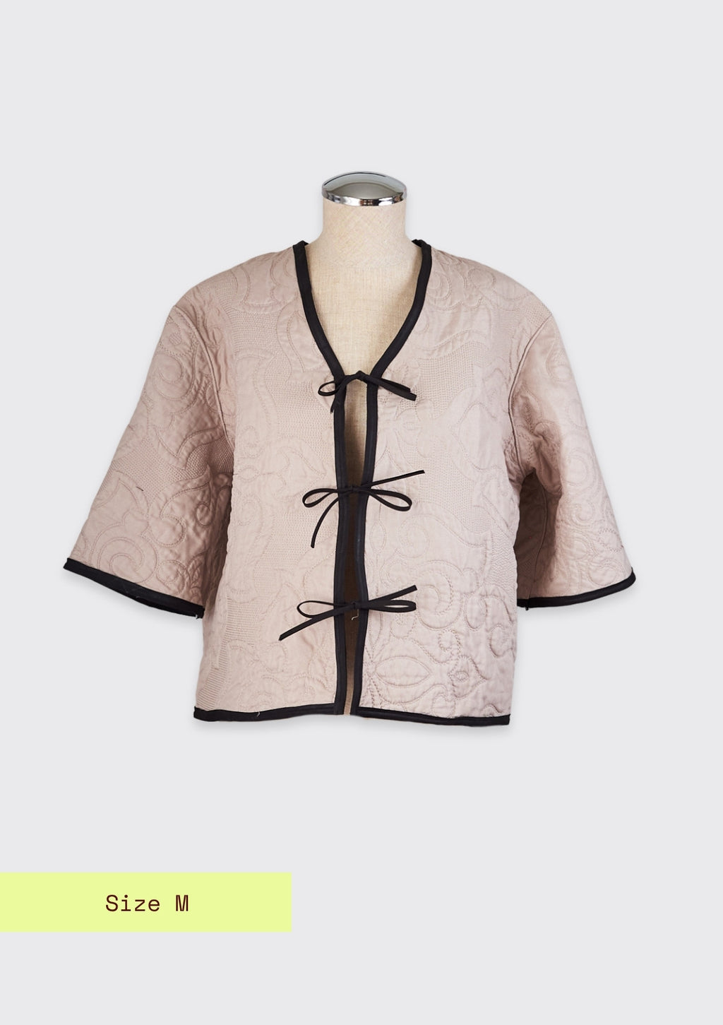 Beige quilted jacket with black trim and tie front closure upcycled from comforter on a mannequin against a light gray background