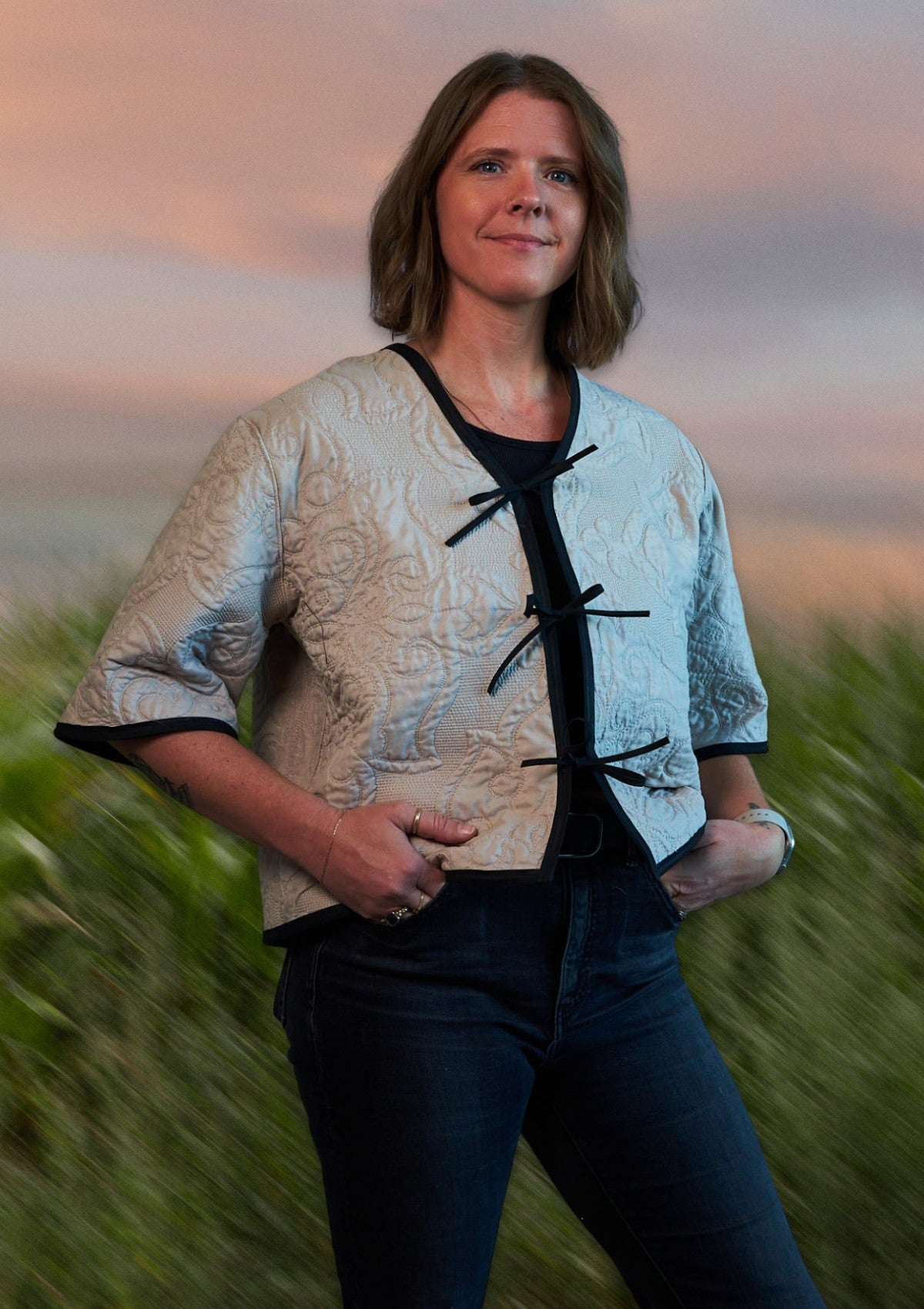 Woman standing in a field with a blurred background wearing a beige patterned tie front jacket upcycled from comforter. 