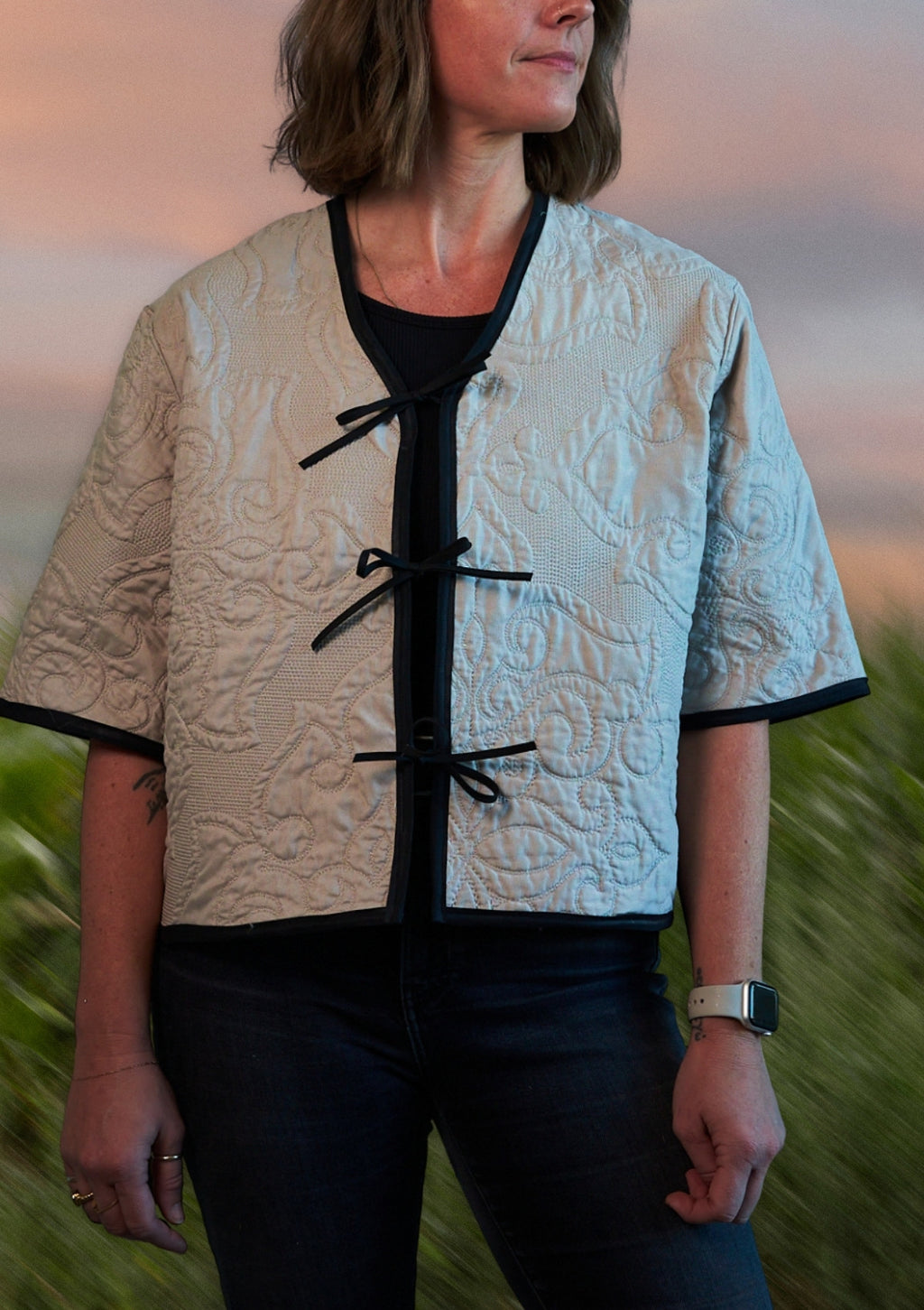 Woman wearing a patterned beige jacket with black tie front closure, upcycled from comforter in a natural setting