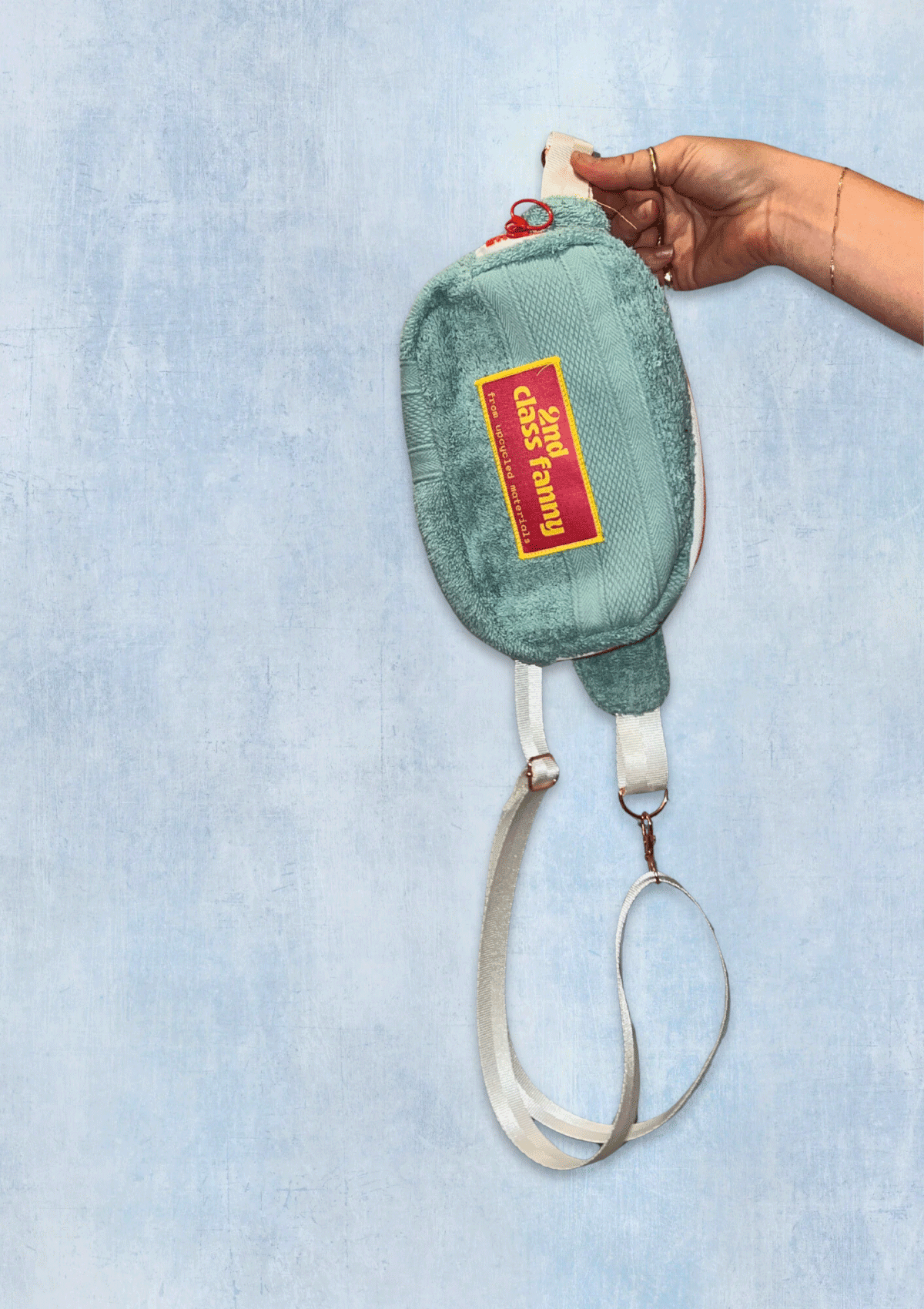 Fanny pack upcycled from teal towel with a pink and yellow label reads 2nd Class Fanny held by a hand against a light blue denim background