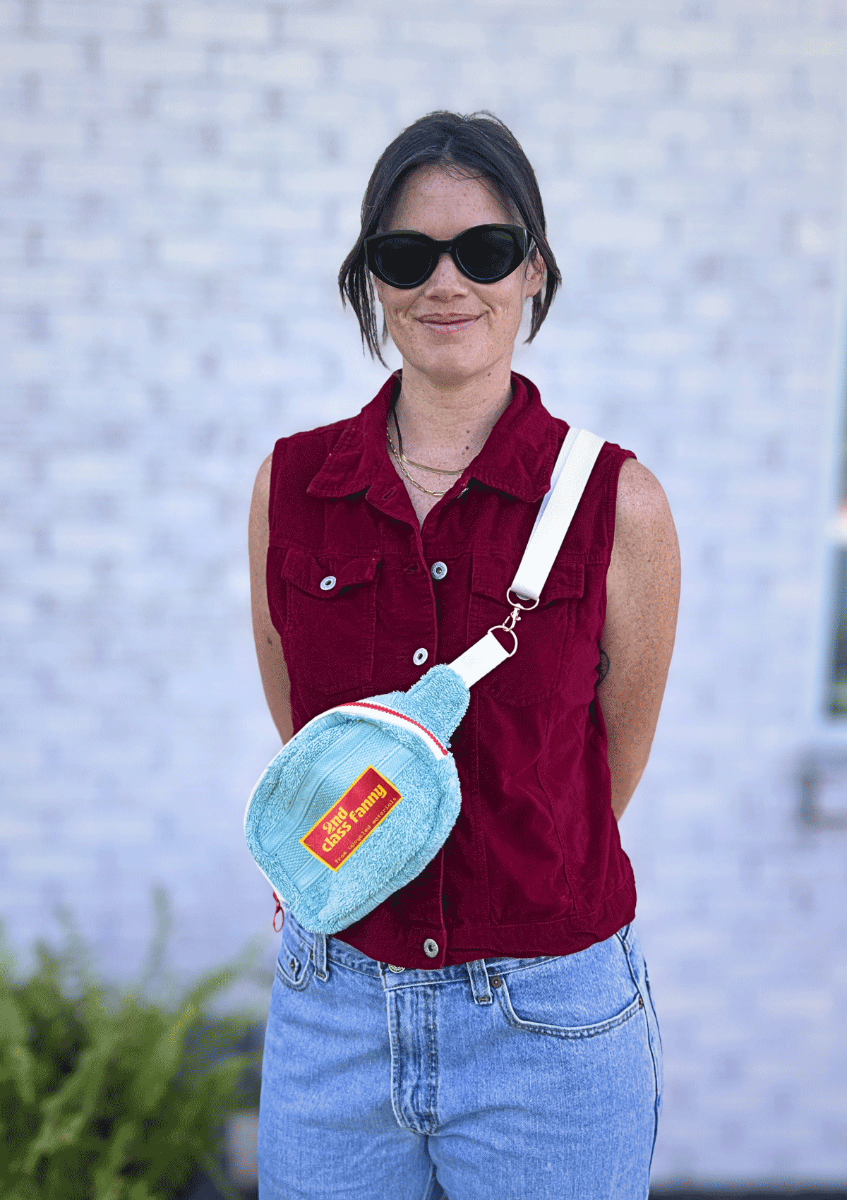 Woman wearing a red sleeveless shirt and blue jeans, holding a Light blue fanny pack upcycled from a towel with a white strap and pink label reads 2nd Class Fanny 