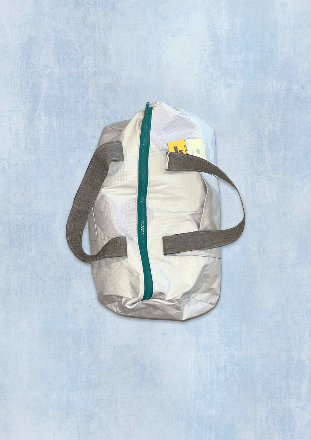 White duffle bag with green zipper and gray straps on a light blue textured background made from upcycled billboard vinyl.