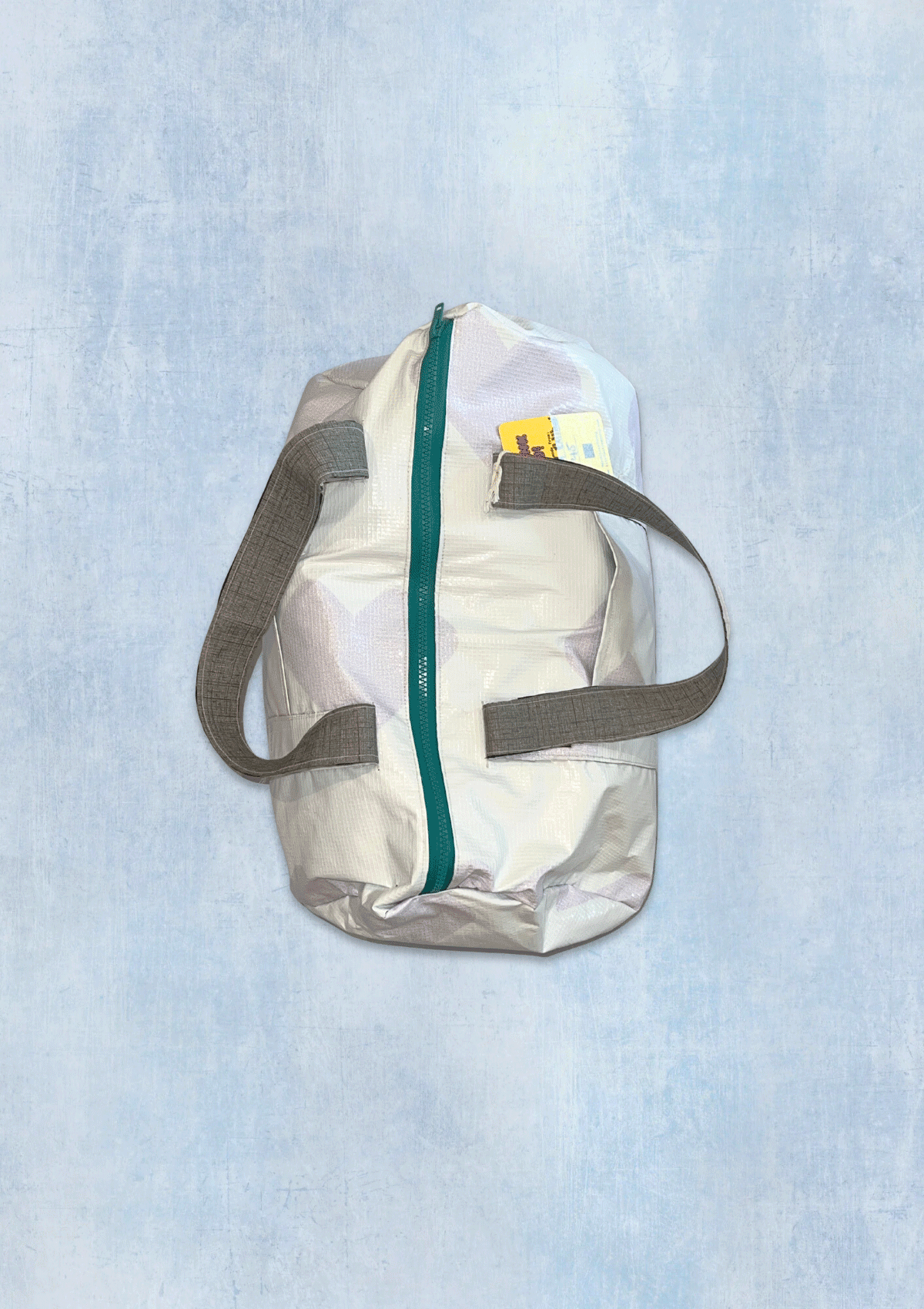 White duffle bag with green zipper and gray straps on a light blue textured background made from upcycled billboard vinyl.