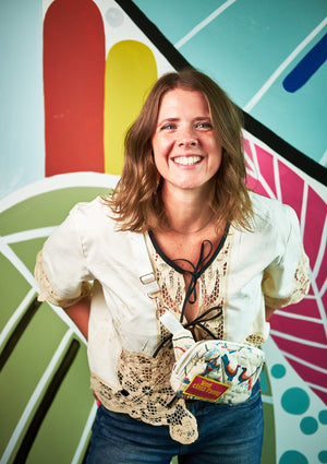 Woman wearing beige lace top with black ties upcycled from a tablecloth with a crossbody quilted bag in front of a colorful abstract wall.
