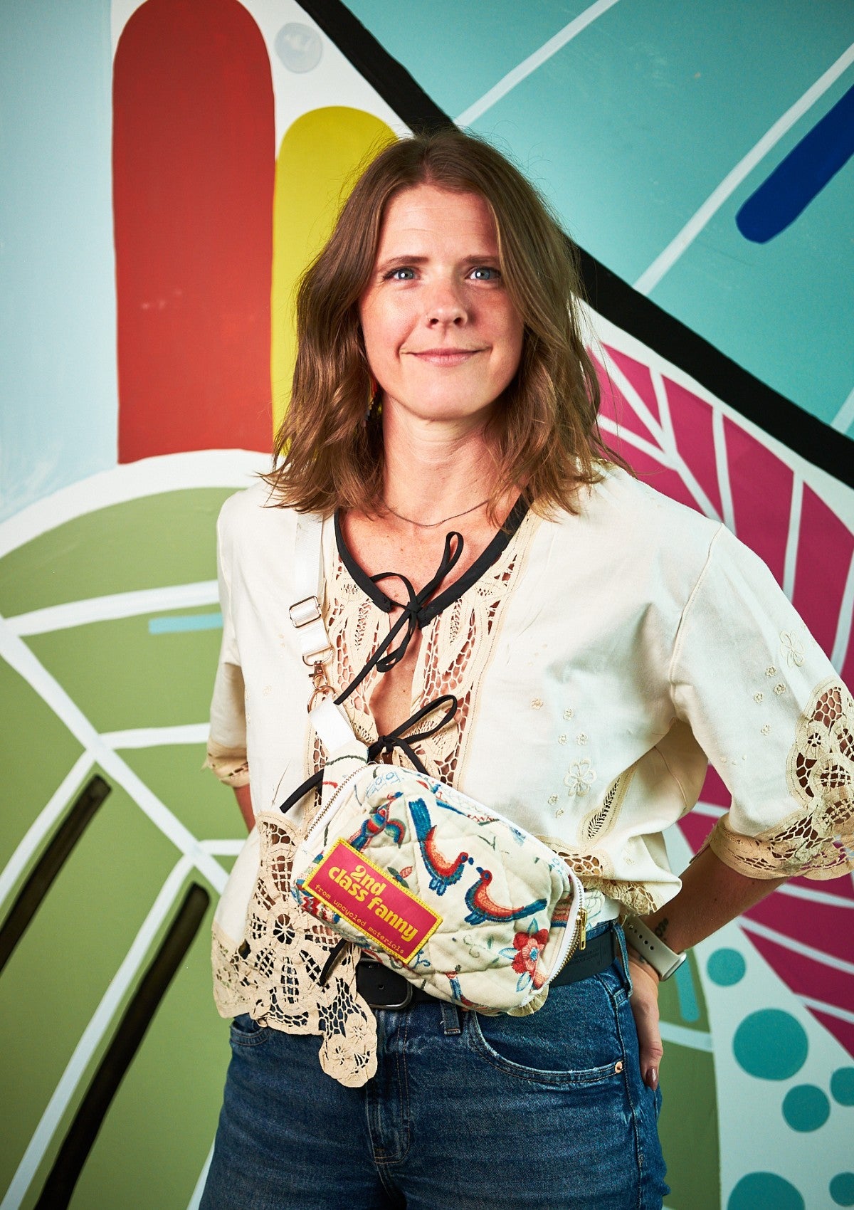 Woman  wearing a beige lace top with black ties holding a colorful bag in front of a multicolored abstract wall.