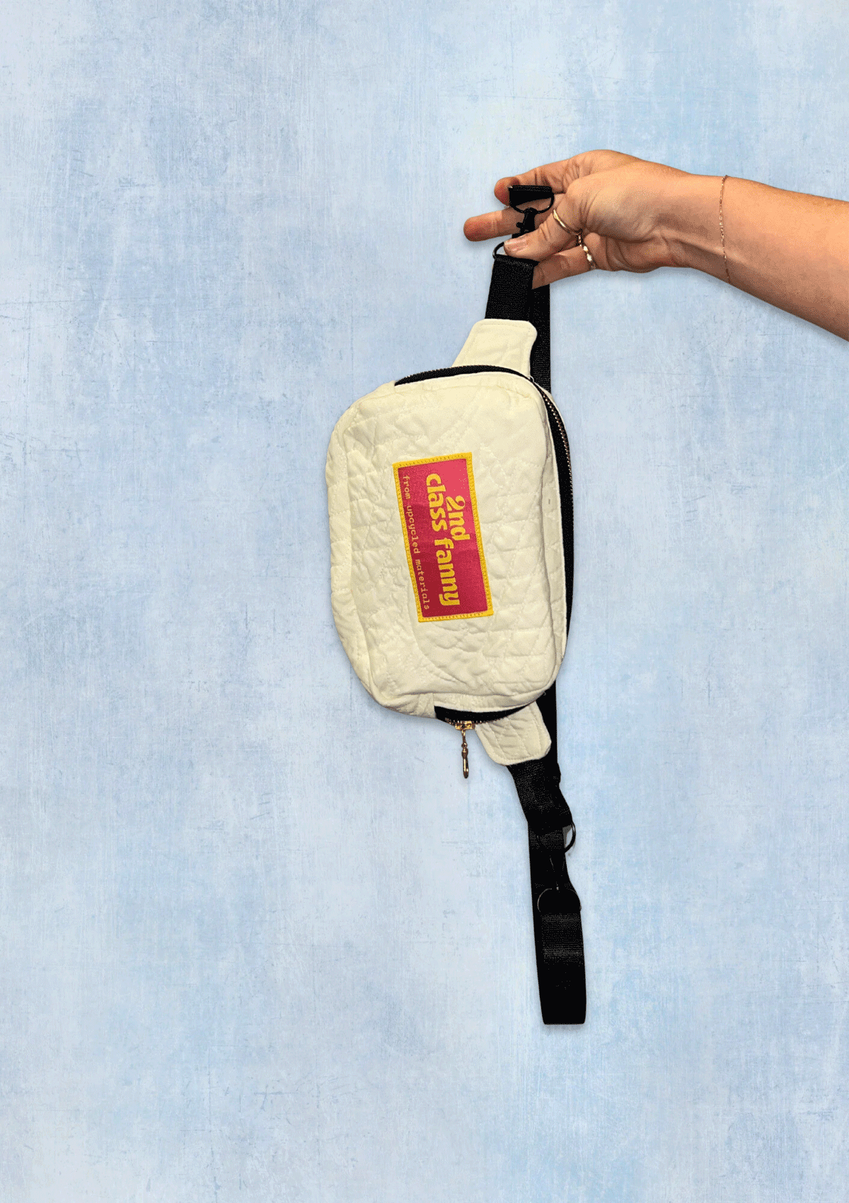 White Fanny Pack, waist bag with a pink label "2nd Class Fanny" held by a hand against a light blue denim background.
