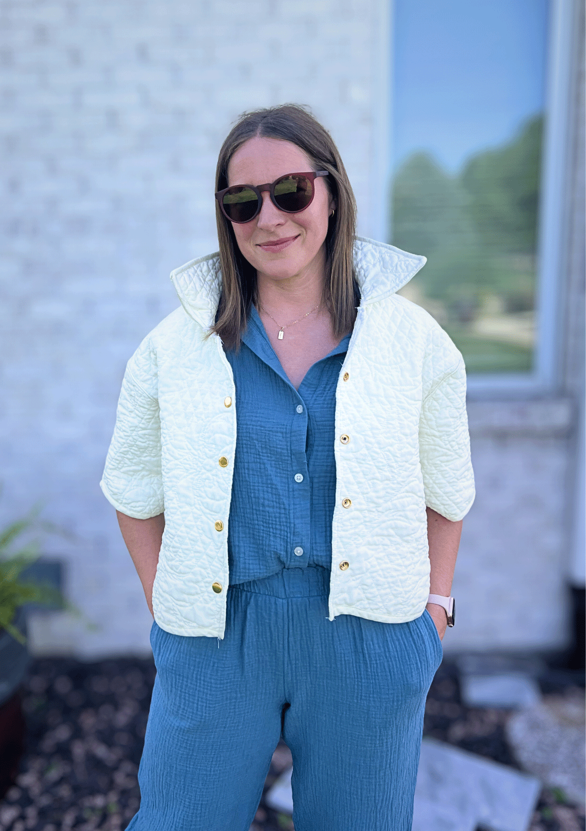 Woman wearing a white jacket upcycled from a quilt over a blue outfit with sunglasses, standing outdoors.