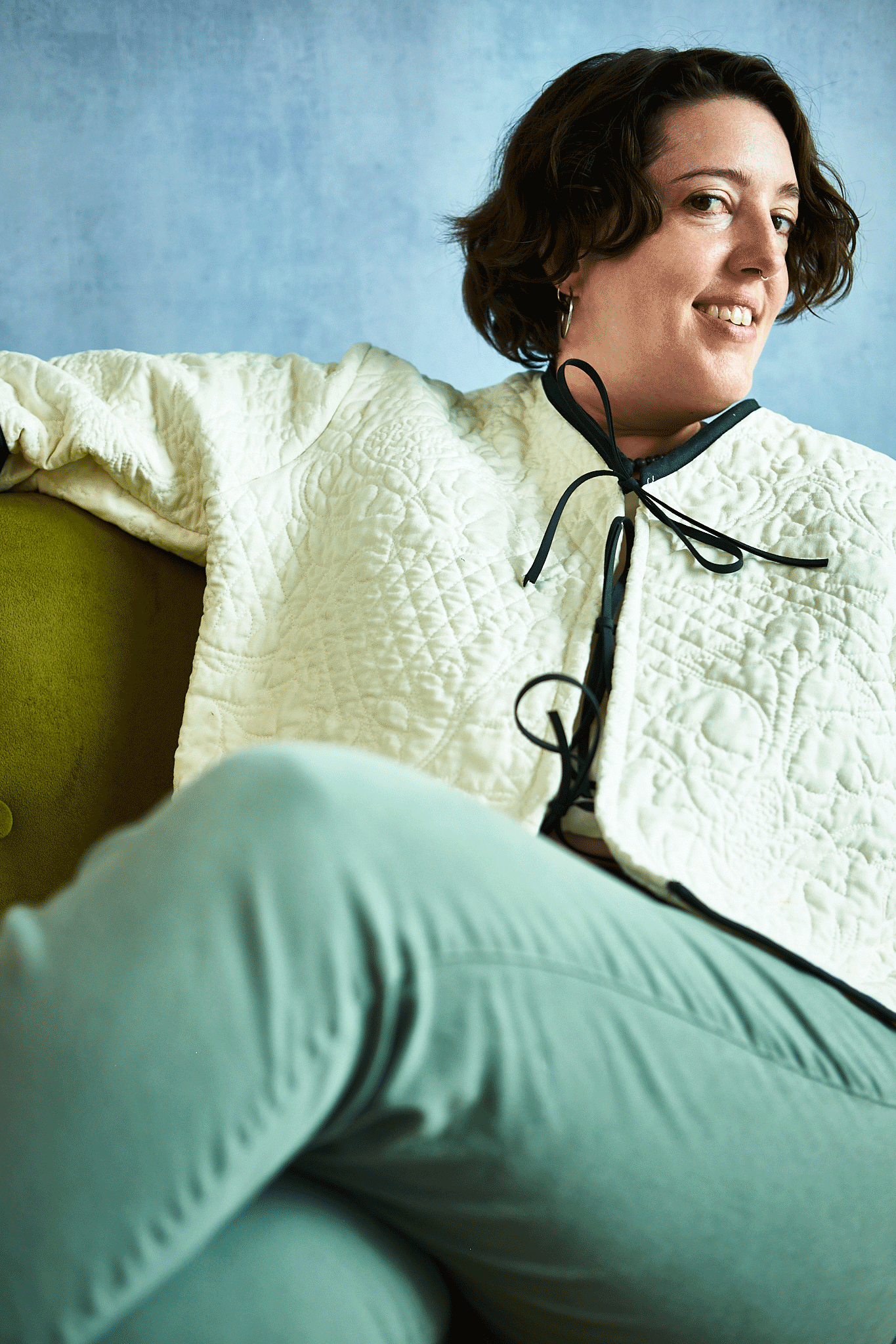 Woman sitting on a green chair wearing a white textured top upcycled from a quilt and light blue pants against a light blue background