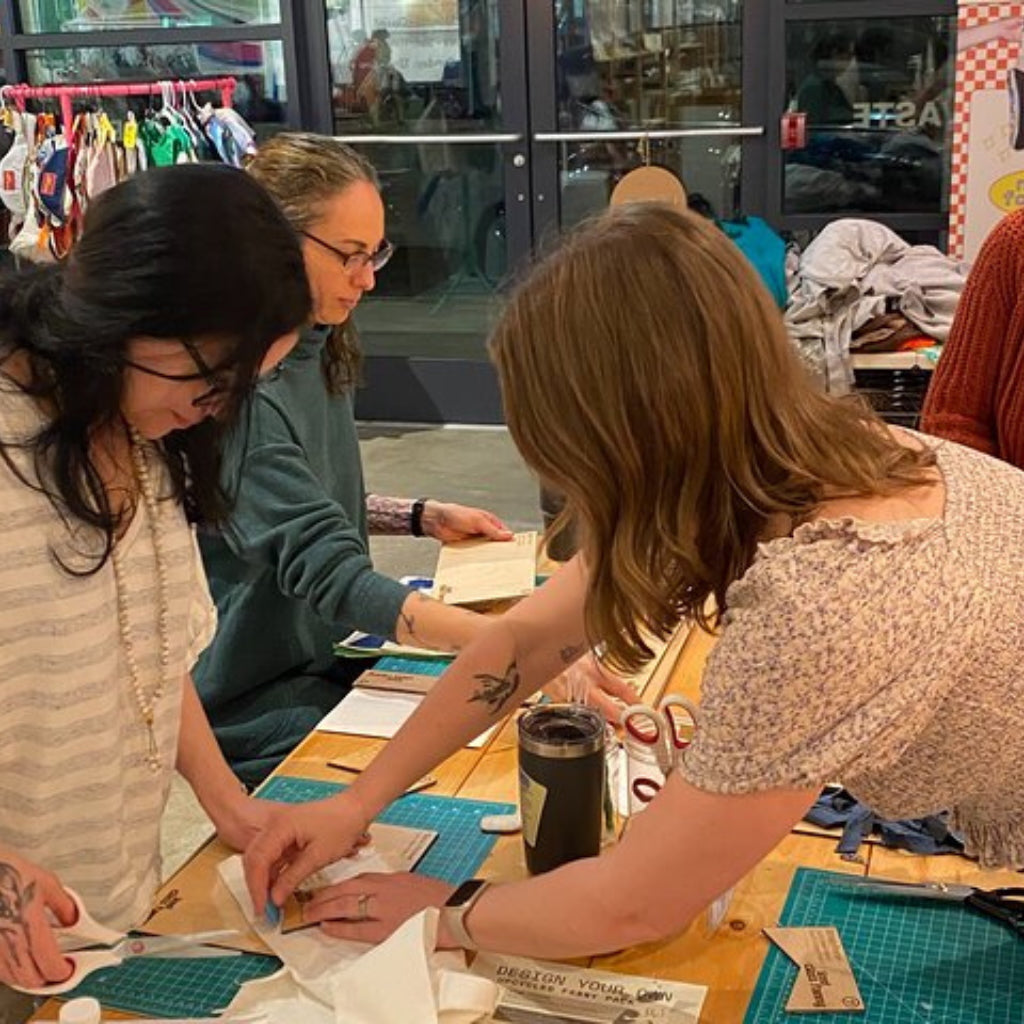 Three women working together at a table with sewing materials in a store setting.